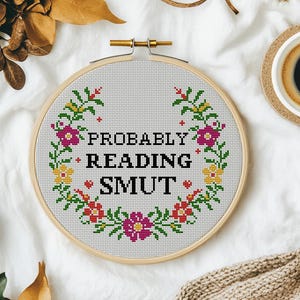 May include: A cross-stitch embroidery hoop with the text "PROBABLY READING SMUT" in black. The text is surrounded by a floral wreath of pink, yellow, and green cross-stitch flowers and leaves. The words "cross-stitch PDF Pattern" are in the bottom left corner.