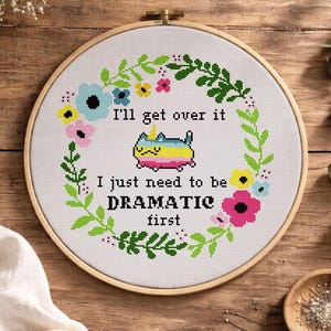 Puede incluir: Un patrón de punto de cruz en un aro de madera, con un diseño de corona floral y el texto "I'll get over it, I just need to be DRAMATIC first." Un gato de dibujos animados con un cuerno de unicornio está en el centro. El texto "CROSS STITCH PATTERN PDF" está en la parte superior.