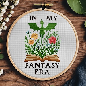May include: Cross-stitch pattern featuring a green dragon flying over a book with flowers and the text "In My Fantasy Era".