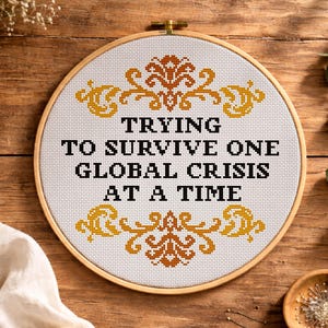 May include: A cross-stitch pattern in a wooden hoop with the text "TRYING TO SURVIVE ONE GLOBAL CRISIS AT A TIME." The design includes decorative gold and brown floral accents. The background is a wooden surface.