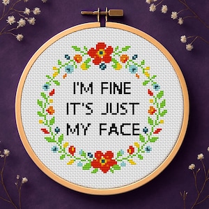 May include: A cross-stitch embroidery hoop with the text "I'M FINE IT'S JUST MY FACE" surrounded by a floral wreath. The embroidery is set against a white background. The words "CROSS-STITCH PATTERN PDF" are at the bottom.