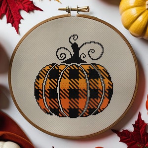Può includere: Modello a punto croce PDF per una zucca a quadri arancioni e neri. Il modello è mostrato in un telaio da ricamo in legno su uno sfondo bianco con decorazioni autunnali.