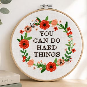 Peut inclure: Un tambour à broder circulaire en bois contenant un motif de point de croix sur tissu blanc. Le motif présente une couronne florale avec des fleurs rouges et pêche et des feuilles vertes entourant le texte noir : YOU CAN DO HARD THINGS.