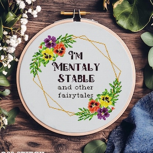 Puede incluir: Un bastidor de bordado de punto de cruz con el texto "I'M MENTALLY STABLE and other fairytales." El diseño presenta un borde floral con flores moradas, amarillas y naranjas. El texto "CROSS-STITCH PDF PATTERN" está en la parte inferior.