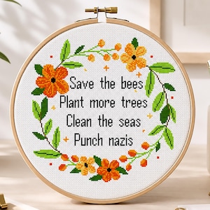 May include: A circular wooden embroidery hoop containing a cross-stitch pattern on white fabric. The design features a floral wreath of orange and yellow flowers with green leaves surrounding the text: Save the bees, Plant more trees, Clean the seas, Punch nazis.
