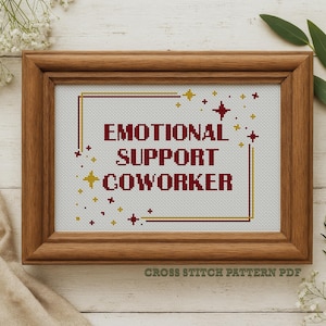 May include: A framed cross-stitch piece with the words "EMOTIONAL SUPPORT COWORKER" in burgundy. The design includes gold and burgundy star accents within a gold and burgundy border. The frame is made of wood.