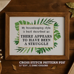 May include: A framed cross-stitch piece with the text "My housekeeping style is best described as: THERE APPEARS TO HAVE BEEN A STRUGGLE." The design is surrounded by green leaf accents. The frame measures 11" x 9".