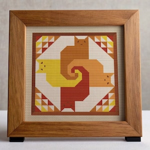 Op de afbeelding: Een ingelijst kruissteek met een geometrisch ontwerp van drie katten in tinten oranje, geel en rood. De katten zijn in een spiraalvorm op een witte achtergrond gerangschikt.