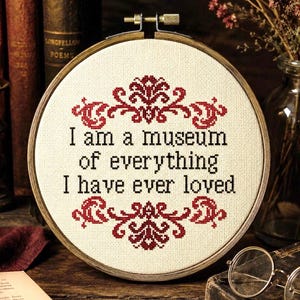 May include: A circular wooden embroidery hoop containing cream-colored fabric with cross-stitched text that reads, "I am a museum of everything I have ever loved," framed by decorative red floral patterns at the top and bottom of the design.