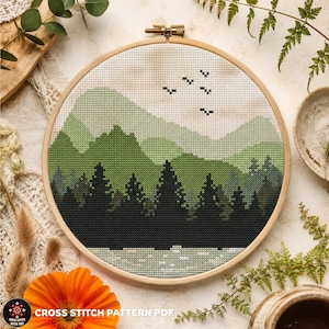 Può includere: Un telaio da ricamo circolare in legno contenente un motivo a punto croce di un paesaggio con pini verde scuro in primo piano, montagne stratificate verde chiaro sullo sfondo e cinque piccoli uccelli neri che volano in un cielo color crema.