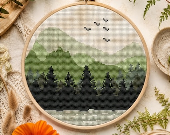 May include: A circular wooden embroidery hoop containing a cross stitch pattern of a landscape with dark green pine trees in the foreground, layered light green mountains in the background, and five small black birds flying in a cream sky.
