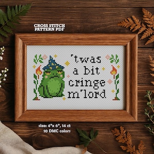 May include: Cross-stitch pattern PDF in a wooden frame. The design features a grumpy green frog wearing a wizard hat, with the text "'twas a bit cringe m'lord." The framed piece measures 10 cm x 15 cm and uses 10 DMC colours.
