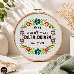 Pode incluir: Um bastidor de bordado circular de madeira contendo tecido Aida branco com um design em ponto cruz. O design apresenta uma coroa de flores em vermelho, azul, roxo e verde ao redor do texto preto que diz: that wasn't very DATA-DRIVEN of you.