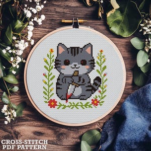 Op de afbeelding: Kruissteek PDF-patroon met een grijze tabby kat met een bloedig mes, omringd door bloemmotieven. De kat zit in een houten borduurring. De tekst "CROSS-STITCH PDF PATTERN" staat onderaan.