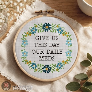 May include: A circular wooden embroidery hoop containing white Aida fabric with a cross-stitch design. The text reads GIVE US THIS DAY OUR DAILY MEDS in black, surrounded by a floral wreath of dark blue, light blue, and yellow flowers.
