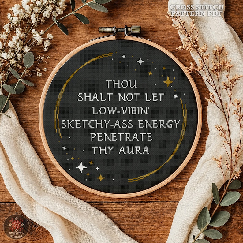 May include: A circular wooden embroidery hoop containing a black fabric cross stitch piece with white text that reads: THOU SHALT NOT LET LOW-VIBIN' SKETCHY-ASS ENERGY PENETRATE THY AURA. The design features a gold stitched crescent and small stars.