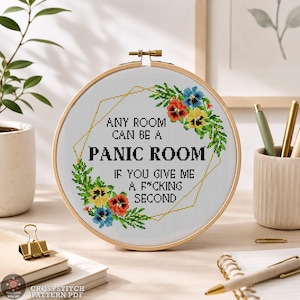 May include: A circular wooden embroidery hoop containing white fabric with a cross-stitch design. The text reads: ANY ROOM CAN BE A PANIC ROOM IF YOU GIVE ME A F*CKING SECOND. It is framed by a geometric gold outline and colorful floral accents.