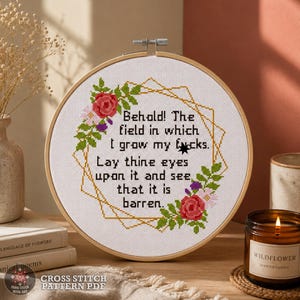 May include: A circular wooden embroidery hoop containing white fabric with a cross-stitch design. The design features a gold geometric frame, red and pink floral accents, and the text: Behold! The field in which I grow my fucks. Lay thine eyes upon it and see that it is barren.