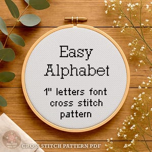 May include: A circular wooden embroidery hoop containing white Aida fabric with black cross-stitched text that reads Easy Alphabet 1" letters font cross stitch pattern. The design is centered within the hoop, which features a metal tightening screw.