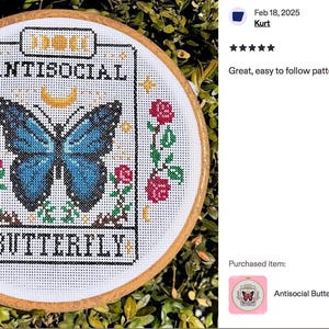 K&ouml;nnte beinhalten: Kreuzstich-Stickerei in einem Holzreifen. Das Design zeigt einen blauen Schmetterling mit den Worten "ANTISOCIAL BUTTERFLY" in schwarzem Text. Rote Rosen und gr&uuml;ne Bl&auml;tter umgeben den Schmetterling. Der Hintergrund ist eine gr&uuml;ne, belaubte Pflanze.