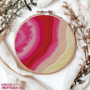 May include: A cross-stitch pattern in a wooden embroidery hoop. The design features a gradient of red, pink, and yellow hues. The text "CROSS STITCH PATTERN PDF" is visible at the bottom. Dried flowers and fabric surround the hoop.