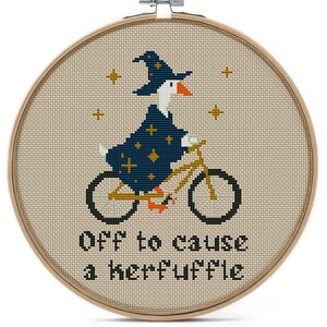 Cross-stitch embroidery in a wooden hoop features a goose riding a bicycle and wearing a wizard hat and robe. The text "Off to cause a kerfuffle" is stitched below the goose. Gold stars are stitched around the goose.