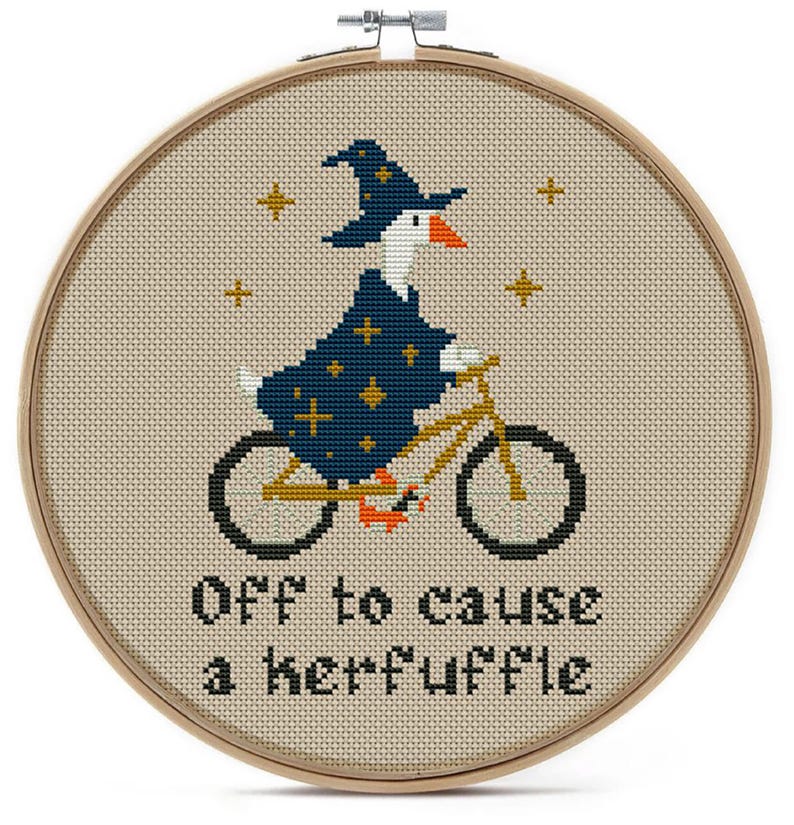 Cross-stitch embroidery in a wooden hoop features a goose riding a bicycle and wearing a wizard hat and robe. The text "Off to cause a kerfuffle" is stitched below the goose. Gold stars are stitched around the goose.