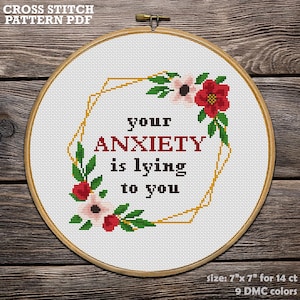 May include: A cross-stitch pattern in a wooden hoop. The design features the text "your ANXIETY is lying to you" surrounded by floral elements and a gold geometric shape. The pattern size is 7" x 7".