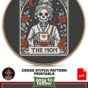 Cross-stitch pattern featuring a skeleton holding a mug with a heart, labeled "THE MOM." The design includes roses, a crescent moon, and stars, framed in a wooden hoop. The pattern is printable.