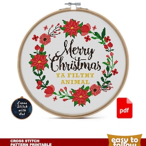 May include: A cross stitch pattern featuring a wreath of red poinsettias and green leaves with the text "Merry Christmas Ya Filthy Animal" in a gold font. The pattern is in a hoop and a PDF download icon is in the corner.