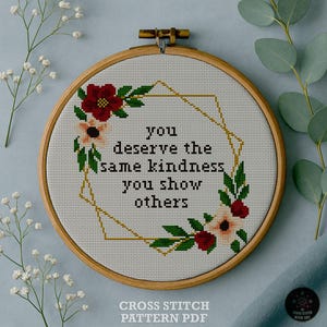 May include: A cross-stitch embroidery in a wooden hoop displays the message "you deserve the same kindness you show others." The design incorporates red and peach floral elements, green leaves, and a gold geometric frame. The text "CROSS STITCH PATTERN PDF" is at the bottom.