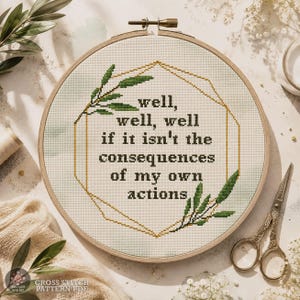 Puede incluir: Un bastidor de bordado circular de madera que contiene tela Aida blanca con un diseño de punto de cruz. El texto dice "well, well, well if it isn't the consequences of my own actions", enmarcado por un borde geométrico dorado y hojas verdes.