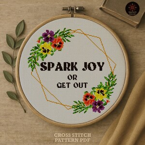 以下が含まれることがあります： 「SPARK JOY OR GET OUT」の文字が入ったクロスステッチ刺繍枠。 金色の幾何学模様と紫、黄色、オレンジ色のカラフルな花がデザインされています。 枠は木製です。