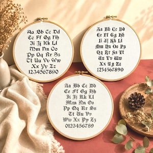 May include: Three circular wooden embroidery hoops containing white fabric with black cross-stitched alphabets and numbers. Each hoop displays the letters A through Z and digits 0 through 9 in a gothic-style font, secured by a metal tension screw.