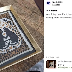 Framed cross-stitch art featuring two black cats with intertwined tails forming a heart, surrounded by floral and heart motifs. The text "THE LOVERS" is at the top. The artwork is in a gold frame.