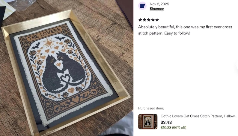 Framed cross-stitch art featuring two black cats with intertwined tails forming a heart, surrounded by floral and heart motifs. The text "THE LOVERS" is at the top. The artwork is in a gold frame.