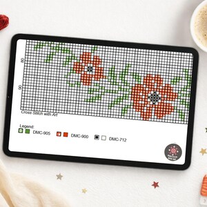 A digital cross-stitch pattern displayed on a tablet, featuring a floral design with red and green elements. The tablet is surrounded by embroidery floss, a cup of coffee, and decorative items. The text "Cross Stitch with Art" is visible.
