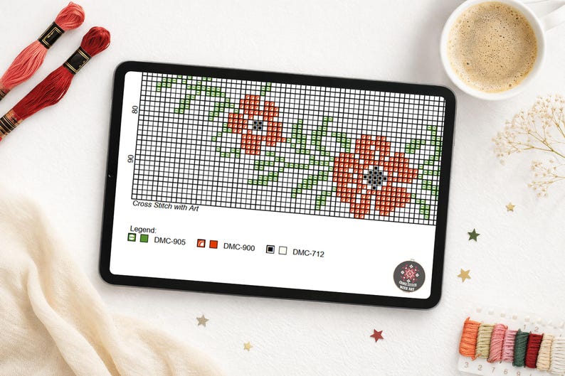 A digital cross-stitch pattern displayed on a tablet, featuring a floral design with red and green elements. The tablet is surrounded by embroidery floss, a cup of coffee, and decorative items. The text "Cross Stitch with Art" is visible.