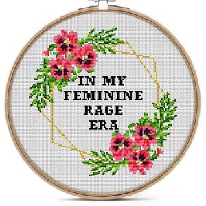 Cross-stitch embroidery in a wooden hoop. The design features the text "IN MY FEMININE RAGE ERA" in black block letters, surrounded by pink flowers and green leaves. A gold geometric shape frames the text.