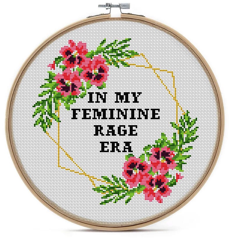 Cross-stitch embroidery in a wooden hoop. The design features the text "IN MY FEMININE RAGE ERA" in black block letters, surrounded by pink flowers and green leaves. A gold geometric shape frames the text.