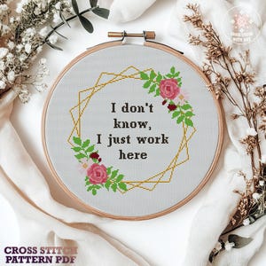 May include: A cross-stitch embroidery hoop with the text "I don't know, I just work here." The design includes a gold geometric frame, pink roses, and green leaves. The words "Cross Stitch Pattern PDF" are at the bottom of the image.