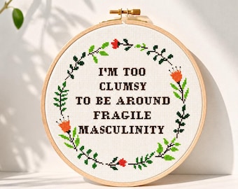 Pode incluir: Um bastidor de bordado circular de madeira contendo tecido branco com um design em ponto cruz. O texto I'M TOO CLUMSY TO BE AROUND FRAGILE MASCULINITY está bordado em letras castanho-escuras, rodeado por uma coroa de folhas verdes e flores laranja.