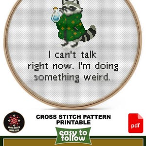 May include: Cross-stitch pattern in a wooden hoop featuring a raccoon wearing a green coat and holding a potion. The text reads, "I can't talk right now. I'm doing something weird." The design size is 6.5" x 6".