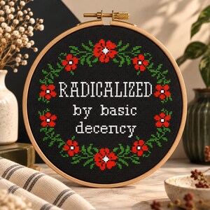 A cross-stitch pattern PDF with the words "RADICALIZED by basic decency" in white. The design is surrounded by a wreath of red flowers and green leaves. The hoop is set against a wooden background with floral accents.