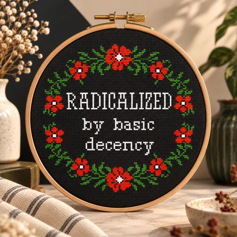 A cross-stitch pattern PDF with the words "RADICALIZED by basic decency" in white. The design is surrounded by a wreath of red flowers and green leaves. The hoop is set against a wooden background with floral accents.