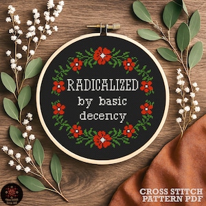 Könnte beinhalten: Ein schwarzer Kreuzstich-Stickrahmen mit dem weißen Schriftzug "RADICALIZED by basic decency". Das Design ist von einem Kranz aus roten Blumen und grünen Blättern umgeben. Der Rahmen liegt auf einem Holzgrund mit floralen Akzenten.
