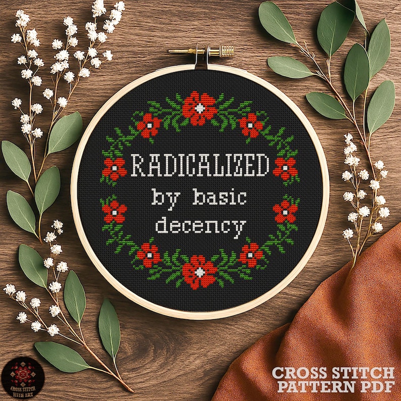 A cross-stitch pattern PDF with the words "RADICALIZED by basic decency" in white. The design is surrounded by a wreath of red flowers and green leaves. The hoop is set against a wooden background with floral accents.