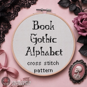 May include: A round wooden embroidery hoop containing white Aida fabric with black cross-stitched text that reads Book Gothic Alphabet cross stitch pattern. The hoop features a metal tightening screw at the top center of the circular frame.