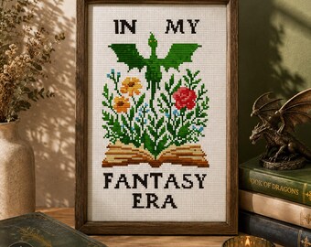 May include: A framed cross-stitch piece on white fabric featuring a green dragon silhouette above an open book with colorful flowers. The text reads IN MY FANTASY ERA in black block letters. The frame is dark brown wood with a simple profile.