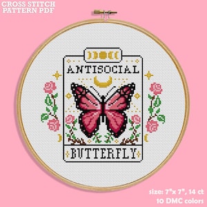 May include: A cross-stitch pattern in a wooden hoop features a pink and black butterfly design with the text "ANTISOCIAL BUTTERFLY." The design is surrounded by pink roses and gold stars. The pattern size is 17.78 cm x 17.78 cm and uses 10 DMC colours.